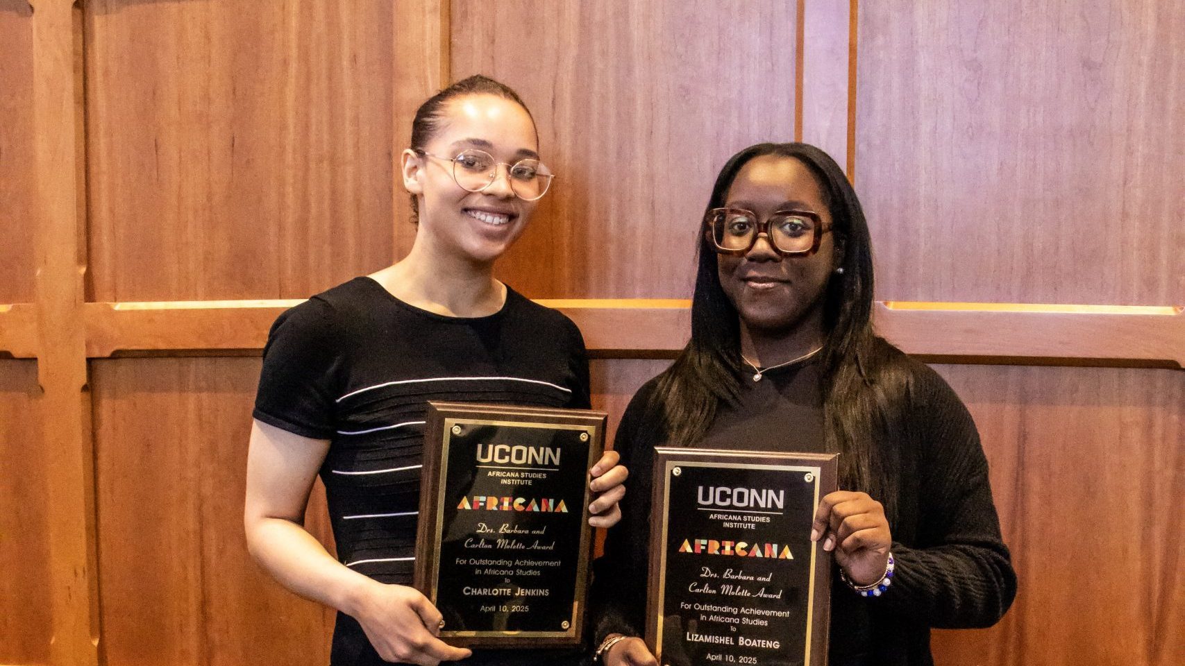 Two Award Recipients stand side by side, smiling and holding UConn Africana Studies awards against a wooden panel backdrop, conveying pride and achievement.
