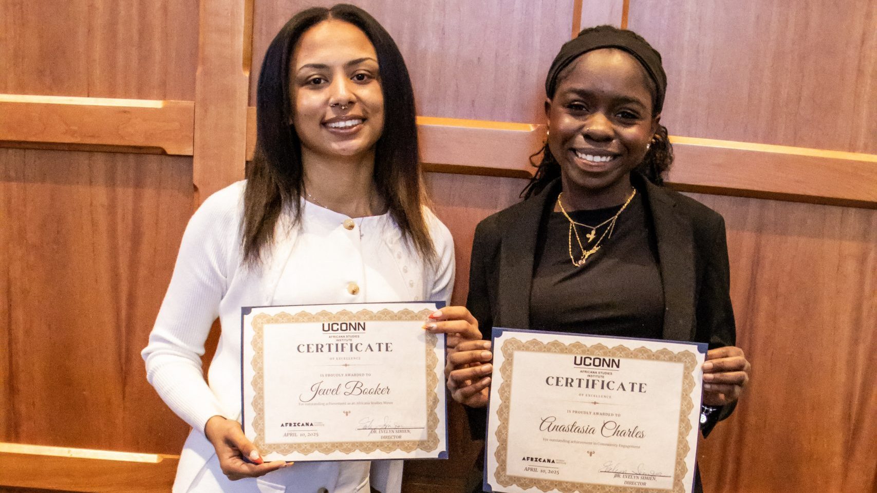 Two Award Recipients smiling and holding certificates against a wooden backdrop. One wears a white dress, the other a black outfit. The mood is celebratory.