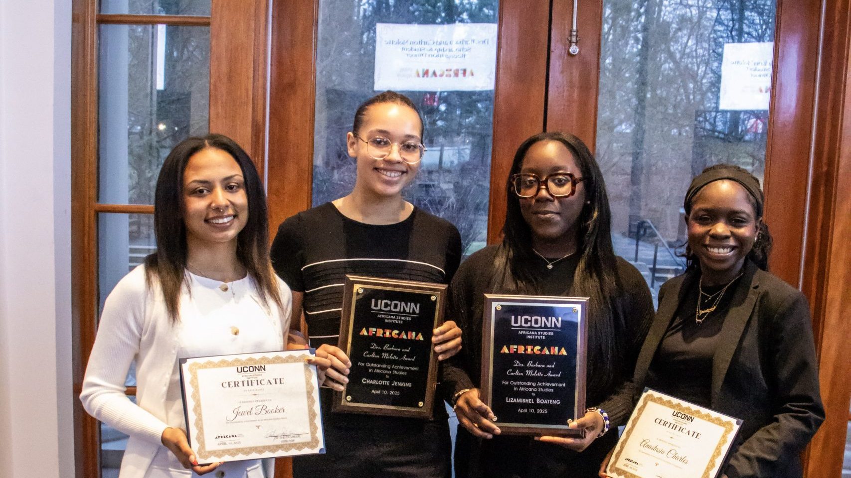 4 award recipients stand smiling indoors holding awards, wearing formal attire. Two display certificates, two hold plaques. A warm, celebratory mood is conveyed.