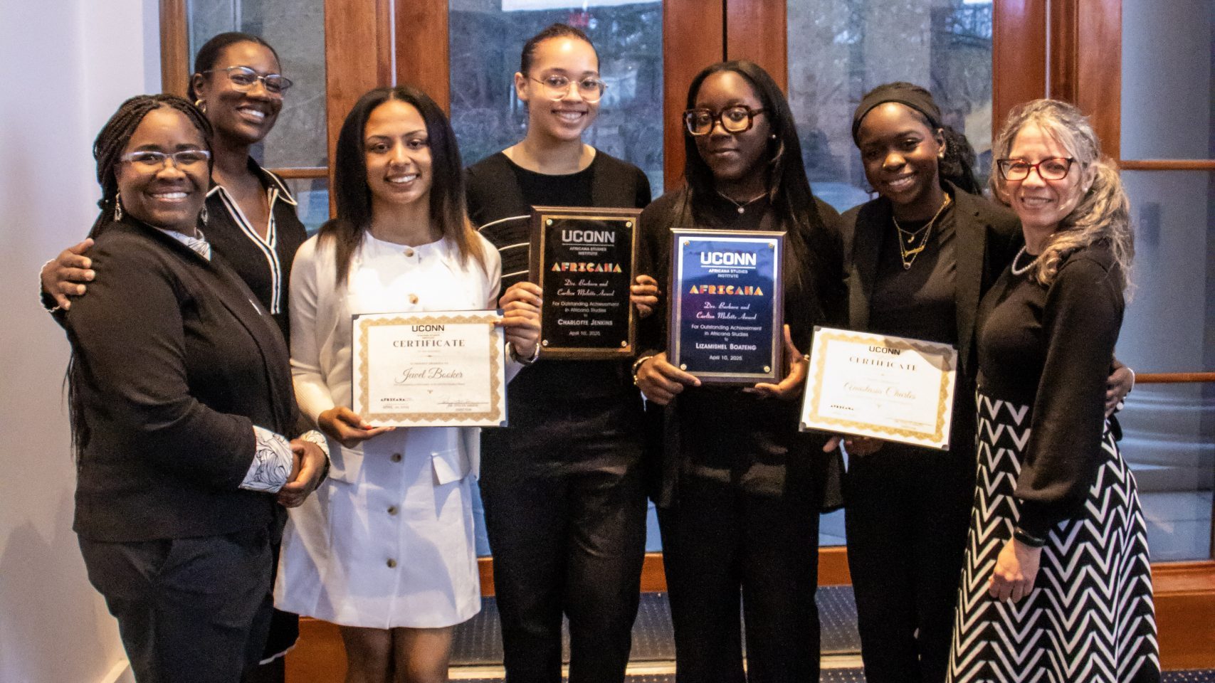 A group of seven people smiling proudly, holding certificates and plaques. They're standing indoors with a glass door behind them, conveying achievement and joy.