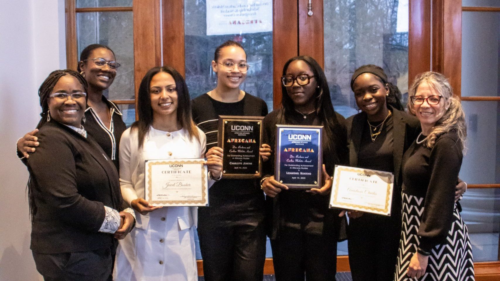 A group of seven people smiling proudly, holding certificates and plaques. They're standing indoors with a glass door behind them, conveying achievement and joy.