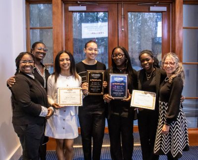 A group of seven people smiling proudly, holding certificates and plaques. They're standing indoors with a glass door behind them, conveying achievement and joy.