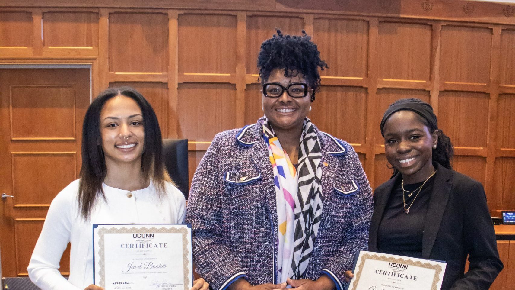 Three women standing in a room with wood paneling. Two smiling women hold certificates. The middle person wears glasses and a patterned jacket.