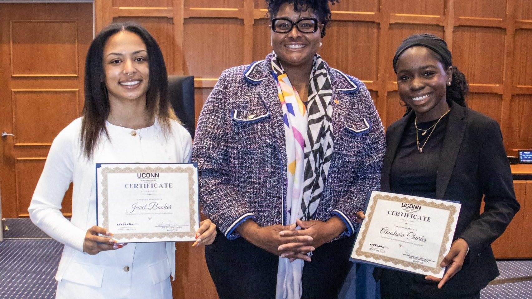 Three women standing in a room with wood paneling. Two smiling women hold certificates. The middle person wears glasses and a patterned jacket.