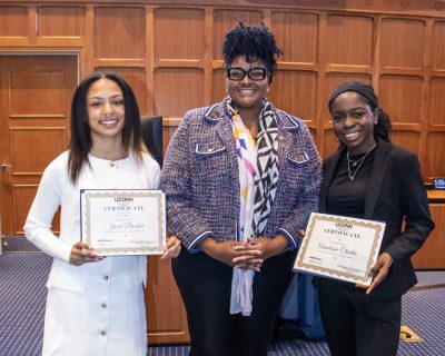 Three women standing in a room with wood paneling. Two smiling women hold certificates. The middle person wears glasses and a patterned jacket.