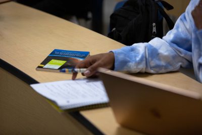 A student in a blue tie-dye sweatshirt takes notes beside a laptop. A book titled "The Autobiography of an Ex-Colored Man" lies nearby on a classroom desk.