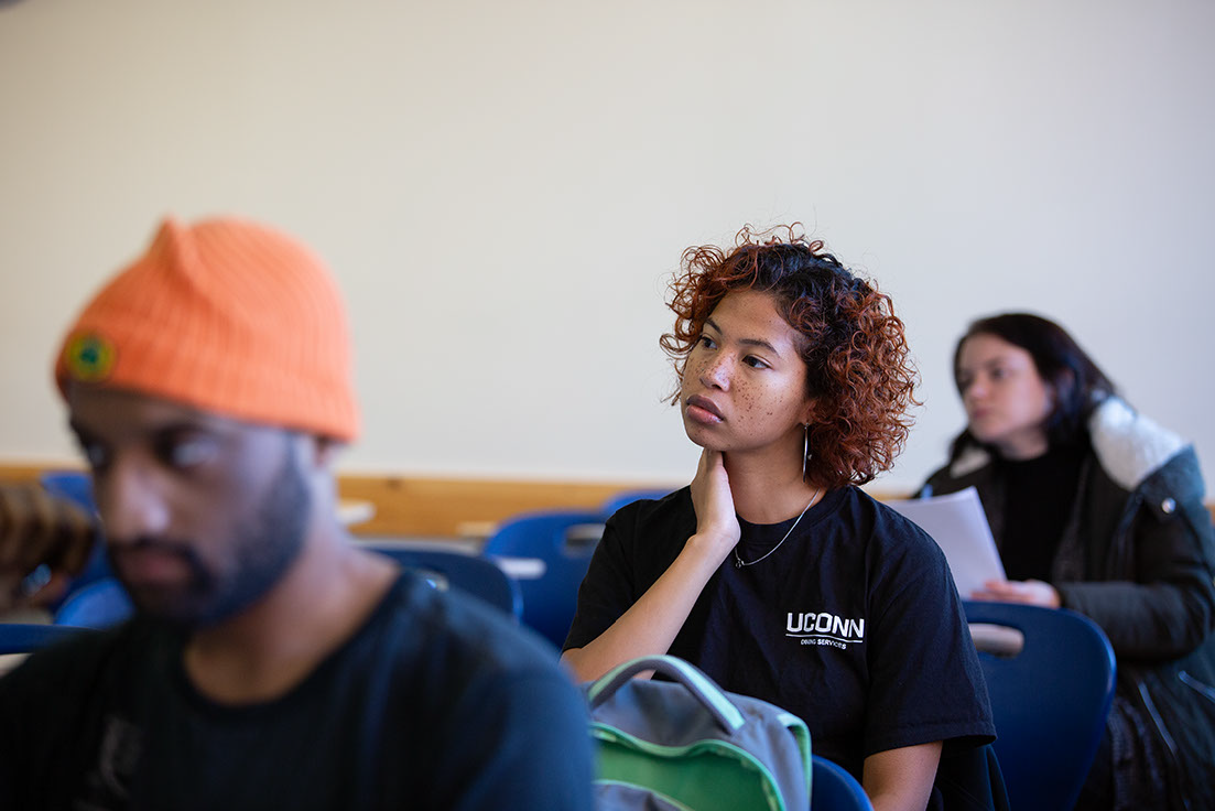A classroom scene with students listening attentively. A woman with curly hair and a pensive expression wears a UConn shirt, conveying focus.