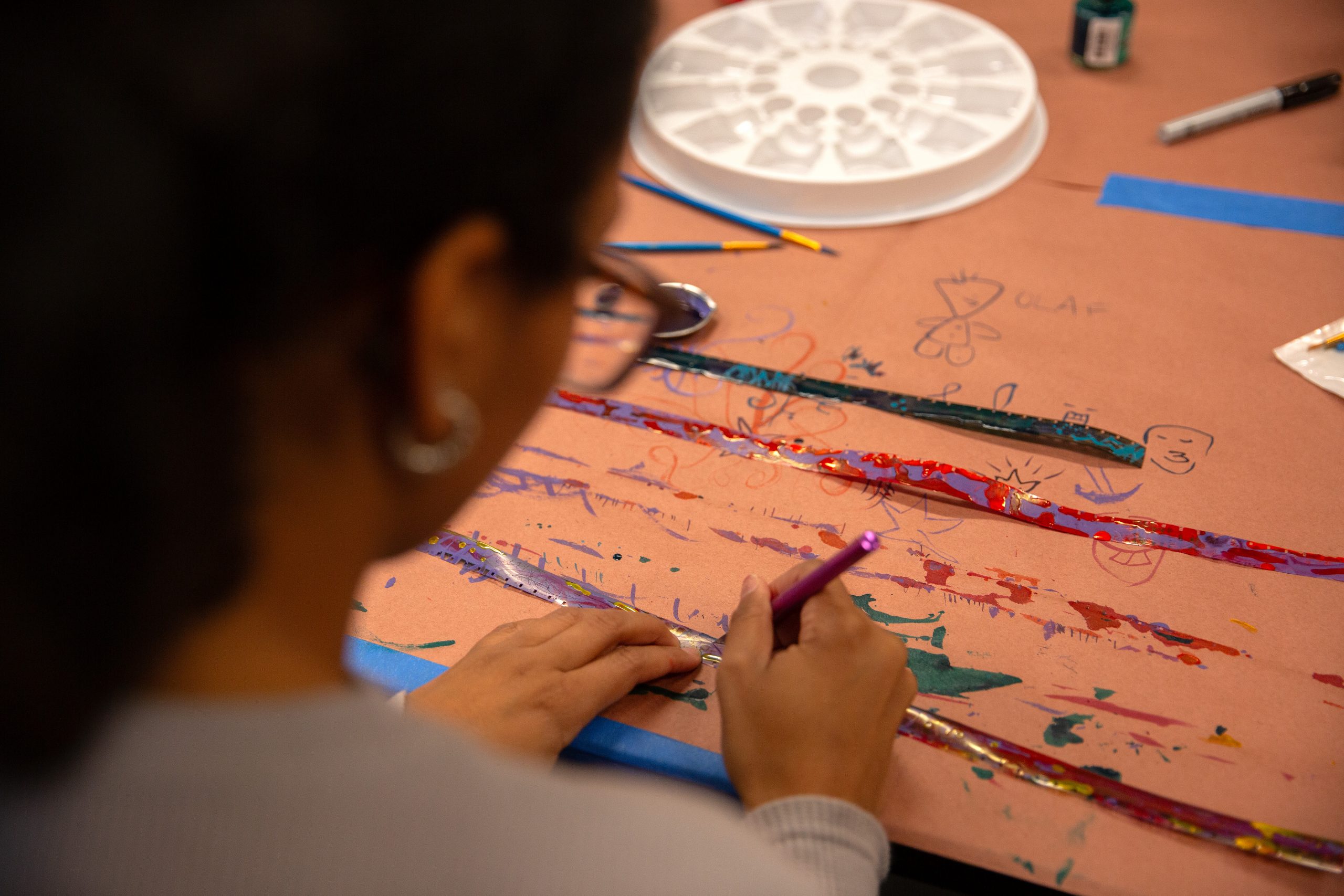 A person focuses on a colorful art project, using a purple pen on paper covered with doodles and paint. The scene is creative and engaging.
