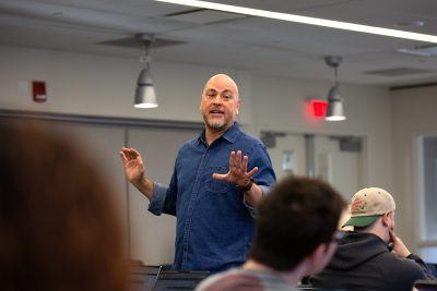 Bryan Cooper Owens in a denim shirt leads a discussion in a classroom. Several students listen, some with laptops open, capturing an engaged learning environment.
