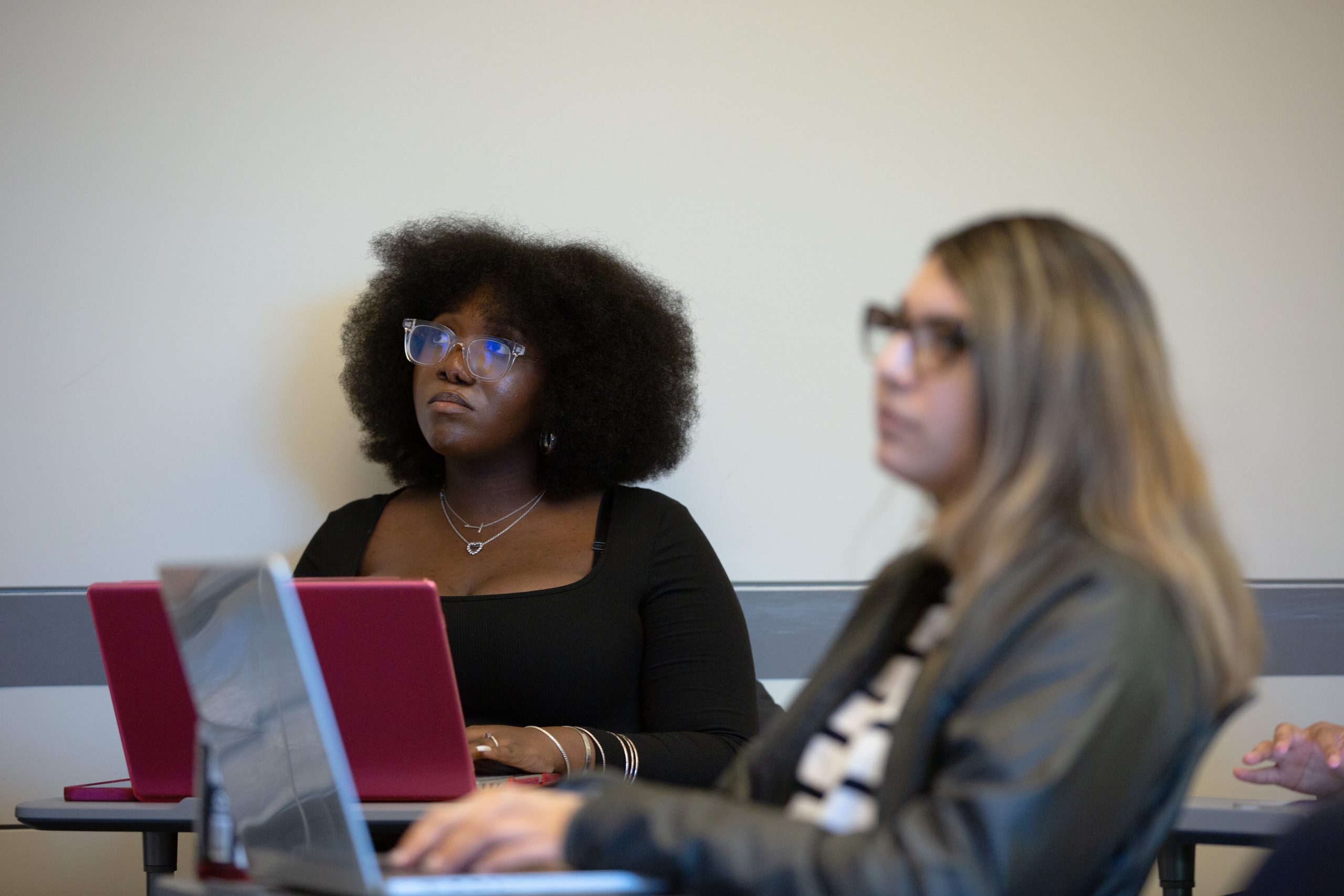 Two students in a classroom, focused and attentive. The student on the left has curly hair and glasses, with a pink laptop. The right student is blurred.