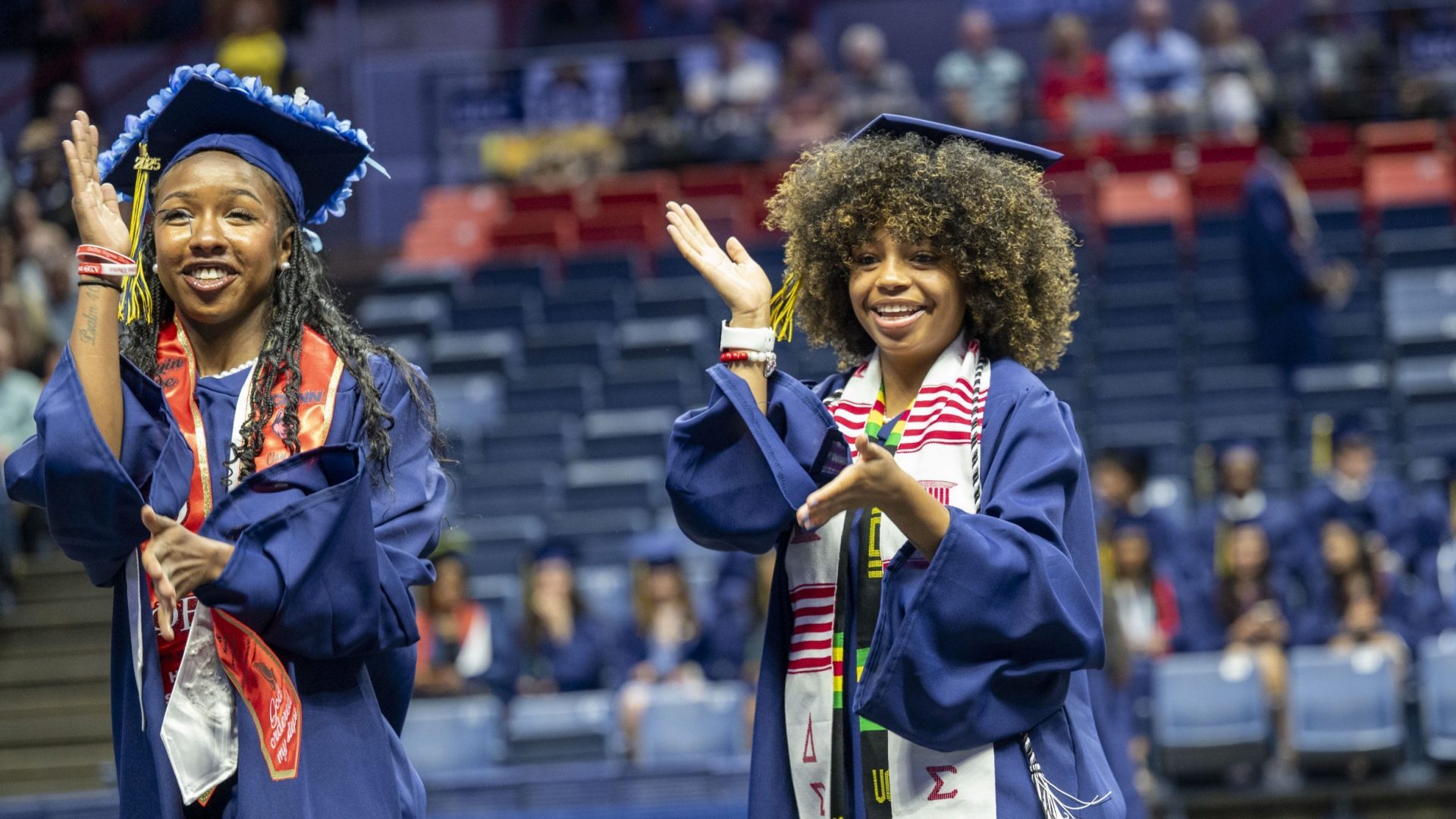 Two graduates in blue gowns and caps applaud joyfully at a ceremony. They wear colorful sashes, standing in a large auditorium with seated attendees.