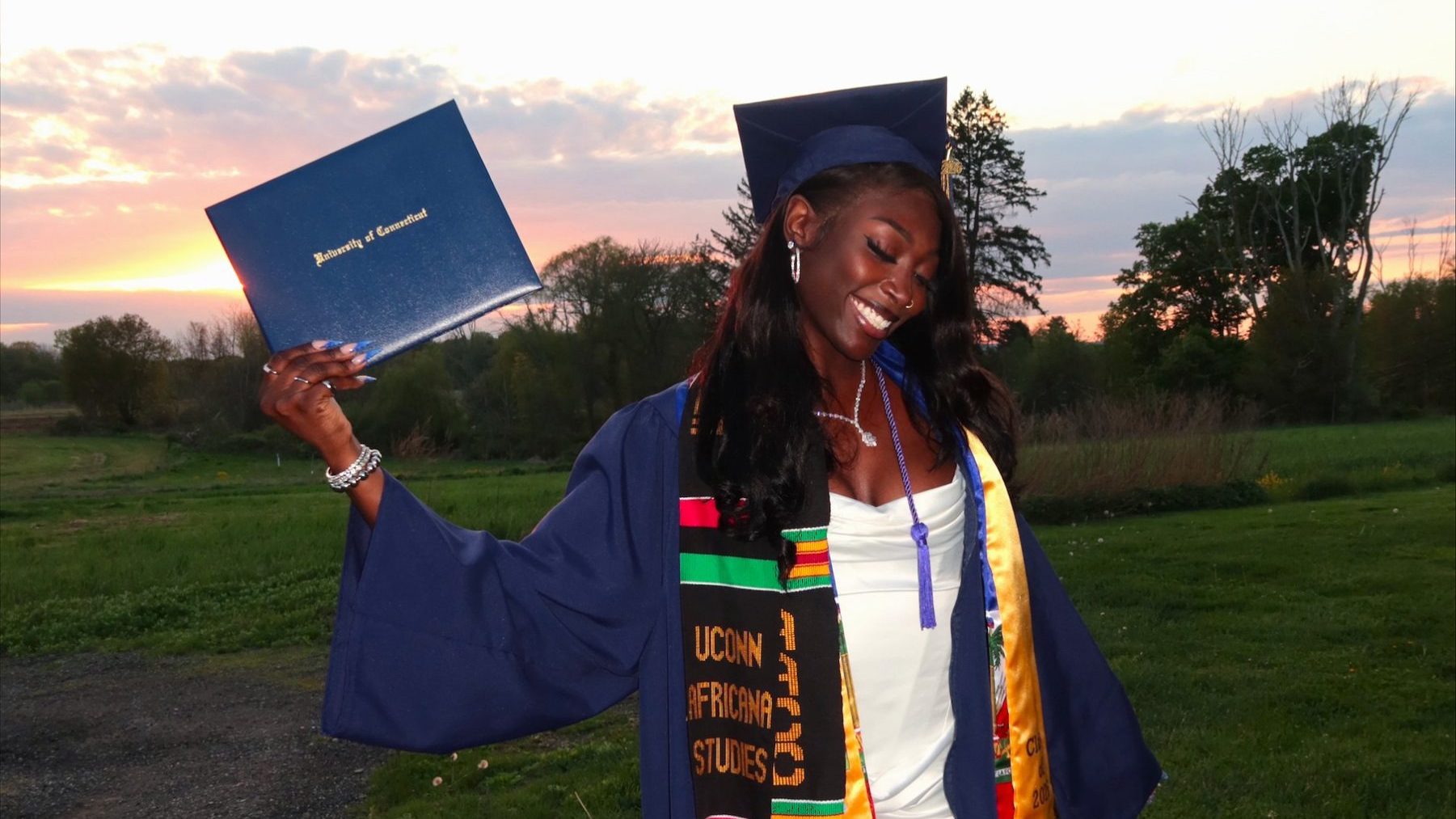 t-pullen-2 A graduate in a navy UConn cap and gown smiles with eyes closed at sunset, holding up a blue "University of Connecticut" diploma cover. She wears a black, gold, red, and green kente stole reading "UConn Africana Studies" over a white dress, standing on a grassy field with trees and a pink-orange sky behind her