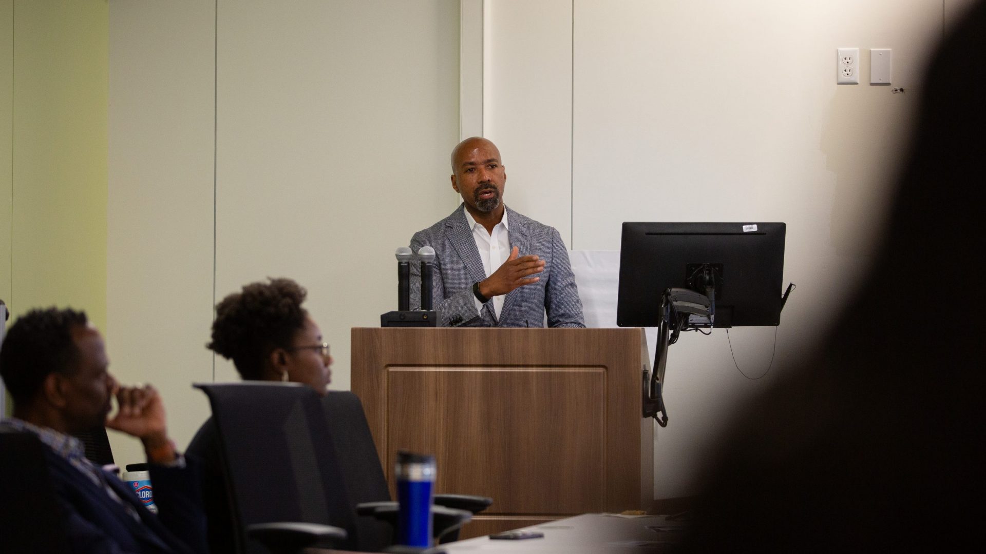A man in a suit speaks at a lectern with a computer and microphones. He gestures to an audience seated in a conference room, conveying a professional tone.