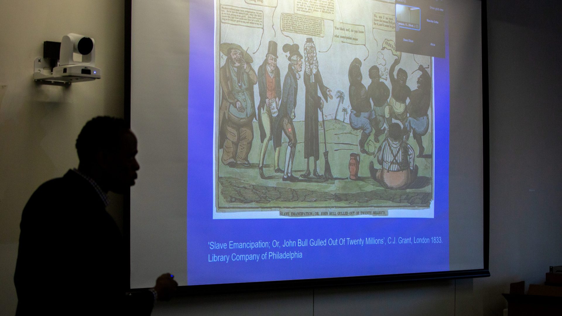 Professor Dexter Gabriel gives a presentation, silhouetted against a projected, colorful cartoon titled "Slave Emancipation." The room is dimly lit, creating a serious tone.