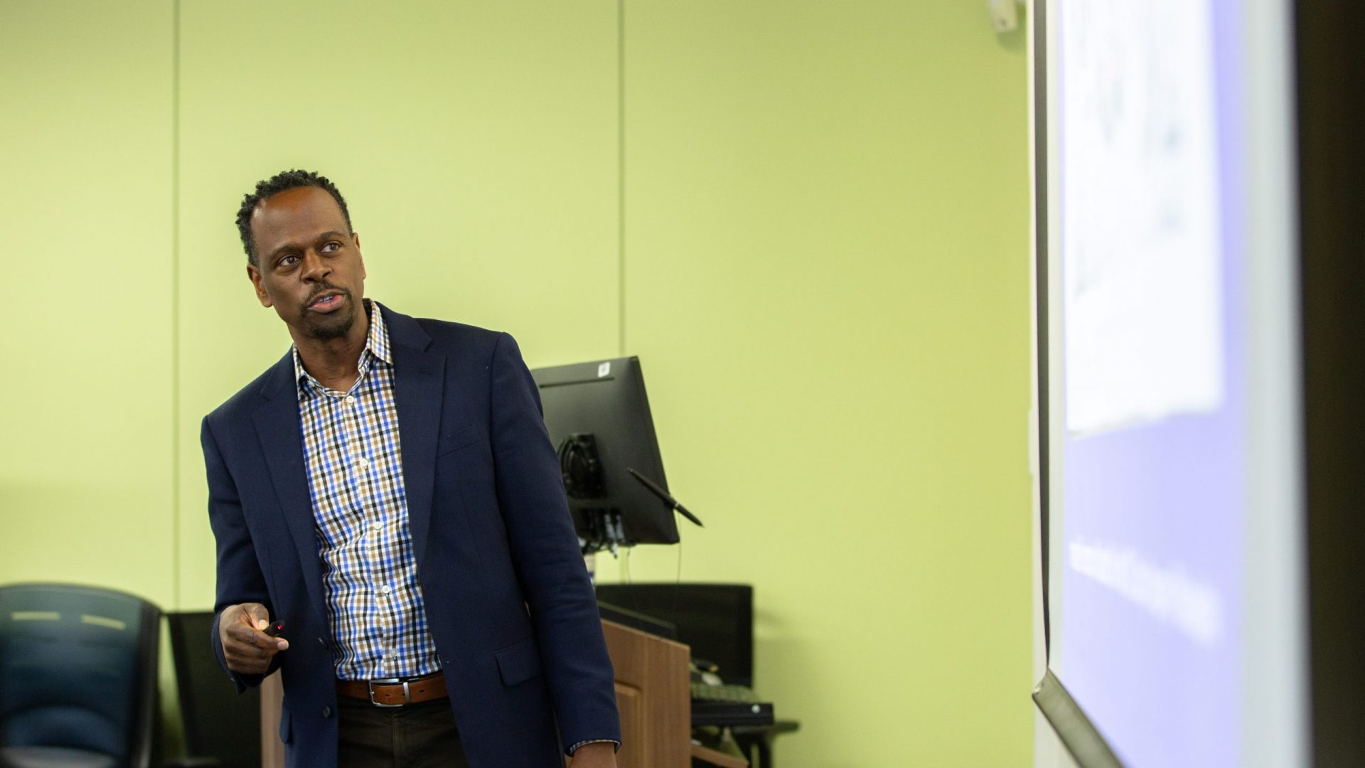 Professor Dexter Gabriel in a blue blazer and checkered shirt giving a presentation, pointing towards a screen. The room has green walls, and a focused, professional atmosphere.