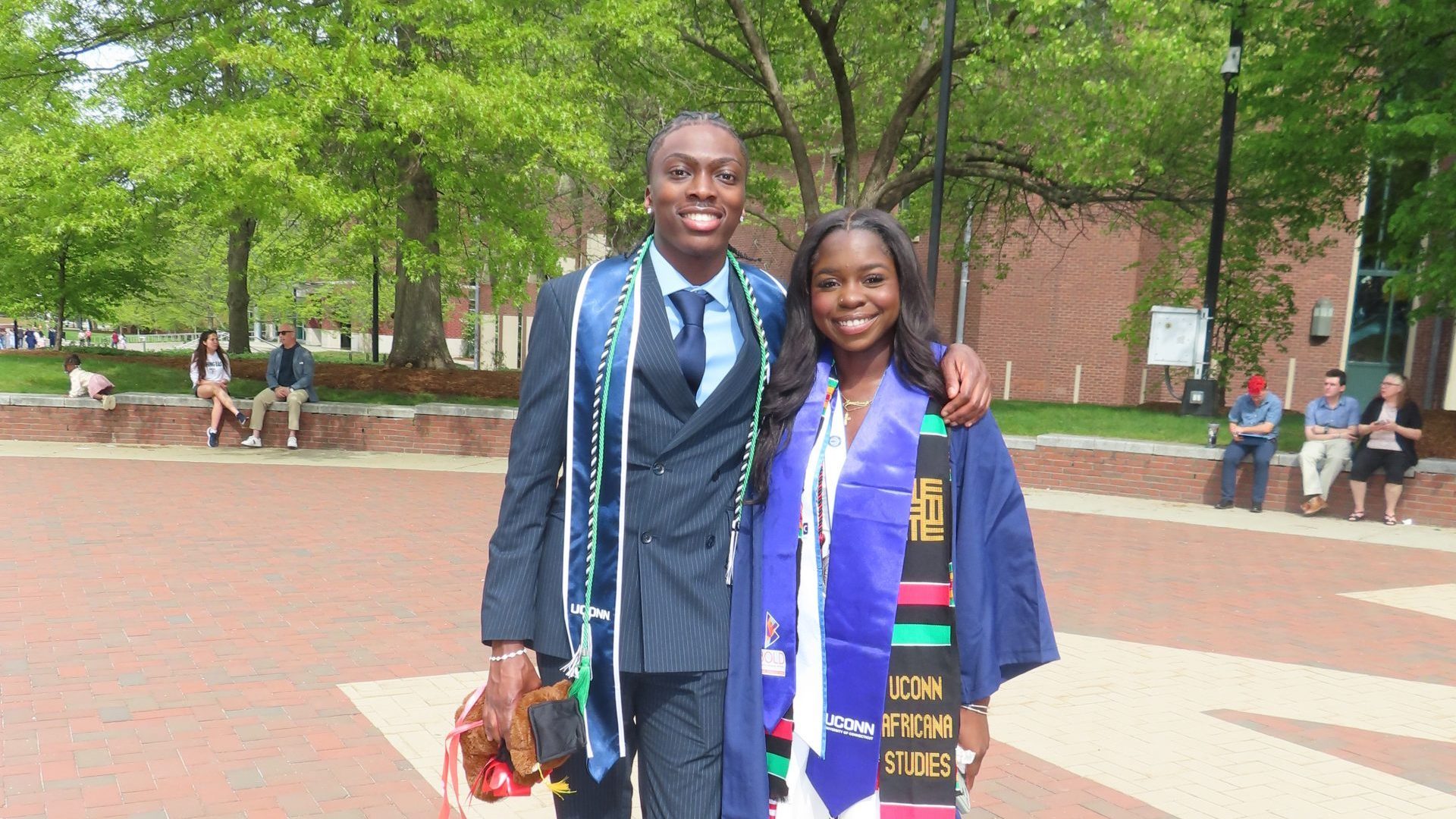 IMG_2582-Commencement 2026 Two graduates pose together on a brick plaza with trees in the background. On the left, a young man in a pinstripe suit and tie wears a UConn stole and honor cords and holds a teddy bear. On the right, a young woman in a navy gown and white dress wears a blue satin stole and a kente stole reading "UConn Africana Studies." Both are smiling at the camera