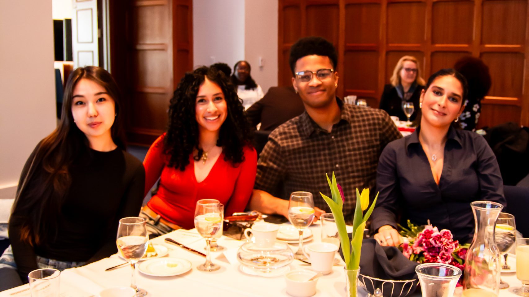 Four guests seated at a formal dinner table smile at the camera during the Molette Dinner. The table is set with white tablecloths, water glasses, coffee cups, a vase of tulips, and a bouquet of pink flowers. A screen reading 'Dinner and Speakers' is visible in the background of the wood-paneled room