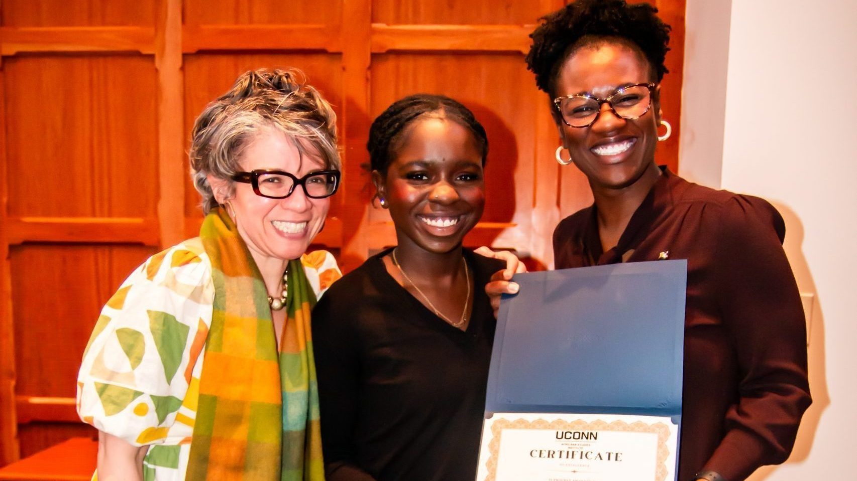 Anastasia Charles, recipient of the Community Engagement Award, smiles between Dr. Evelyn Simien and Amanda Cannada as they hold up her certificate from the UConn Africana Studies Institute. Anastasia wears all black, Dr. Simien wears a white dress with an orange, green, and yellow geometric print and matching scarf, and Amanda wears a dark burgundy dress. They pose in front of wood-paneled walls.