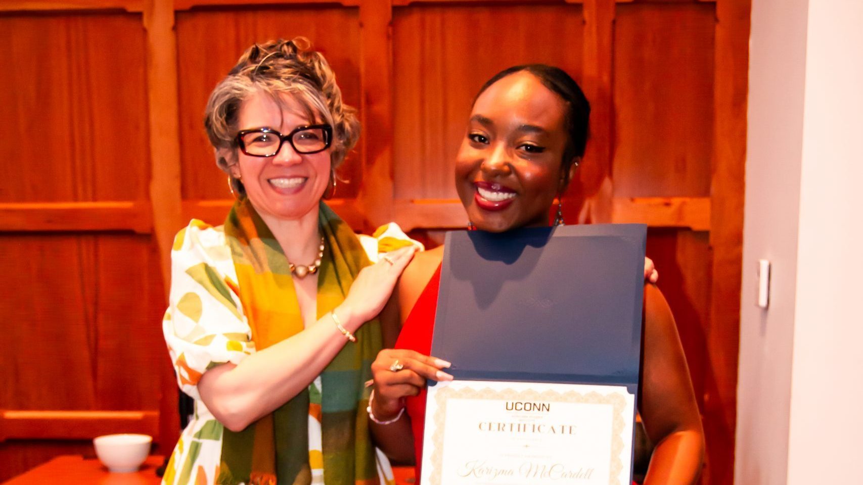 Karizma McCardell, wearing a bright red dress, smiles as she holds up her Academic Excellence in the Minor Award certificate from the UConn Africana Studies Institute. Dr. Evelyn Simien, in a white dress with an orange, green, and yellow geometric print and matching scarf, stands beside her with a congratulatory hand on her shoulder. They pose in front of wood-paneled walls