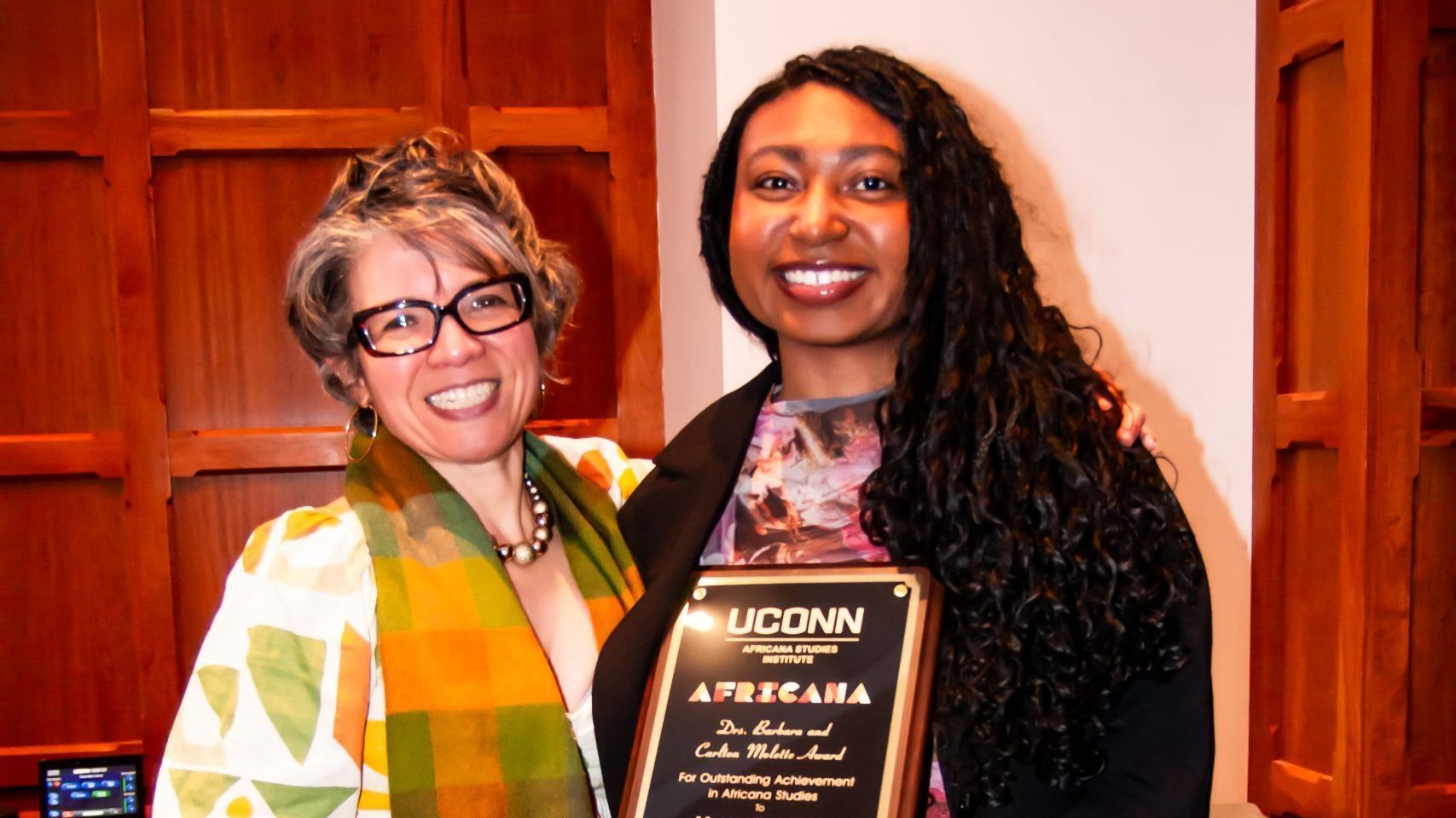 Myeisa Barnhill, recipient of the Drs. Barbara and Carlton Molette Award, smiles as she holds her engraved plaque from the UConn Africana Studies Institute. Beside her, Dr. Evelyn Simien, in a white dress with an orange, green, and yellow geometric print and matching scarf, wraps an arm around her shoulder. They pose in front of wood-paneled walls