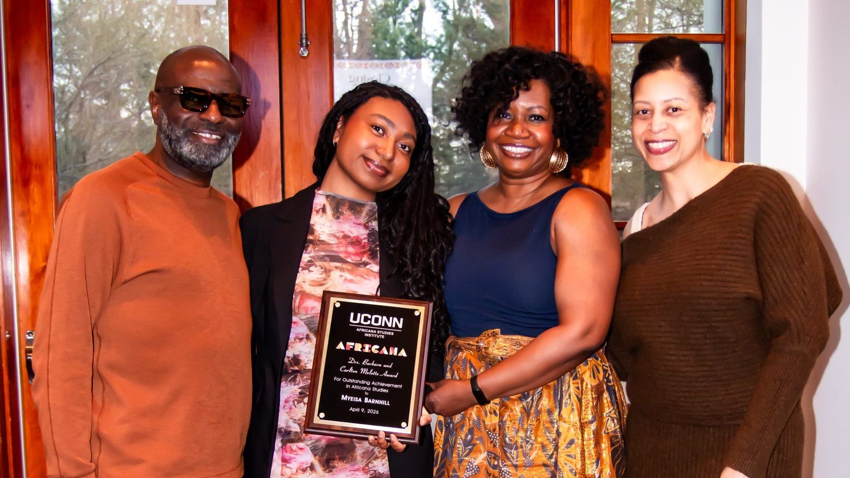 Myeisa Barnhill holds her Drs. Barbara and Carlton Molette Award plaque from the UConn Africana Studies Institute while posing with three others in front of wooden doors with an exit sign. Beside her, Dr. Deirdre Cooper Owens, in a navy top and gold patterned skirt, helps hold the plaque. A man in a rust-colored sweater stands at the far left, and a woman in a brown sweater dress stands at the far right