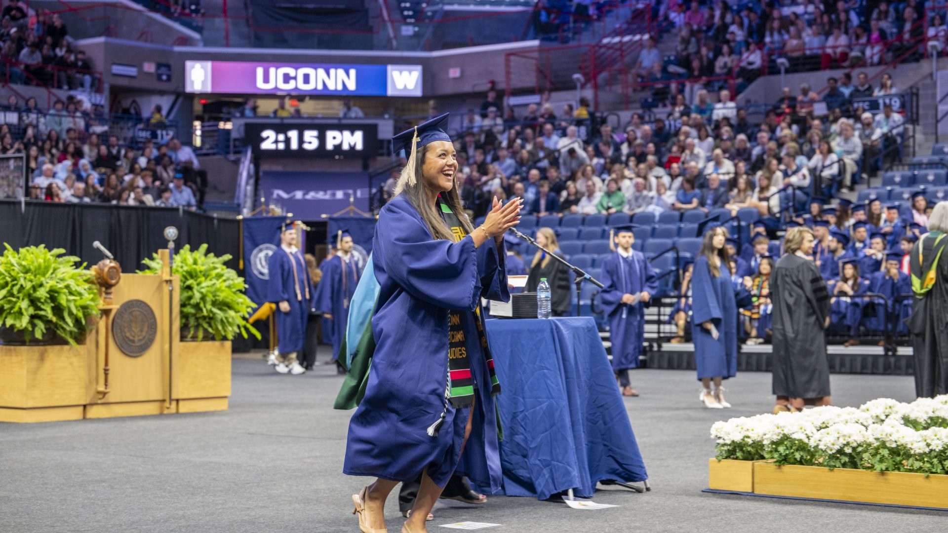 Commencement 2026-business_260509b0283.jpg A graduate in a navy cap and gown claps and smiles as she walks across the floor of UConn's arena during commencement. She wears a UConn Africana Studies kente stole, and the arena scoreboard behind her reads "UCONN 2:15 PM," with the audience filling the stands and other graduates in the background